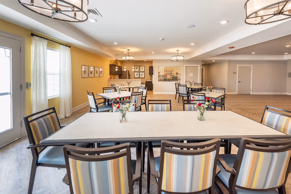 A bright and spacious dining room in a senior living facility with multiple tables and chairs arranged neatly. Each table has a small vase with flowers. The room features yellow and beige walls, large windows with white curtains, modern light fixtures, and a kitchenette area in the background.