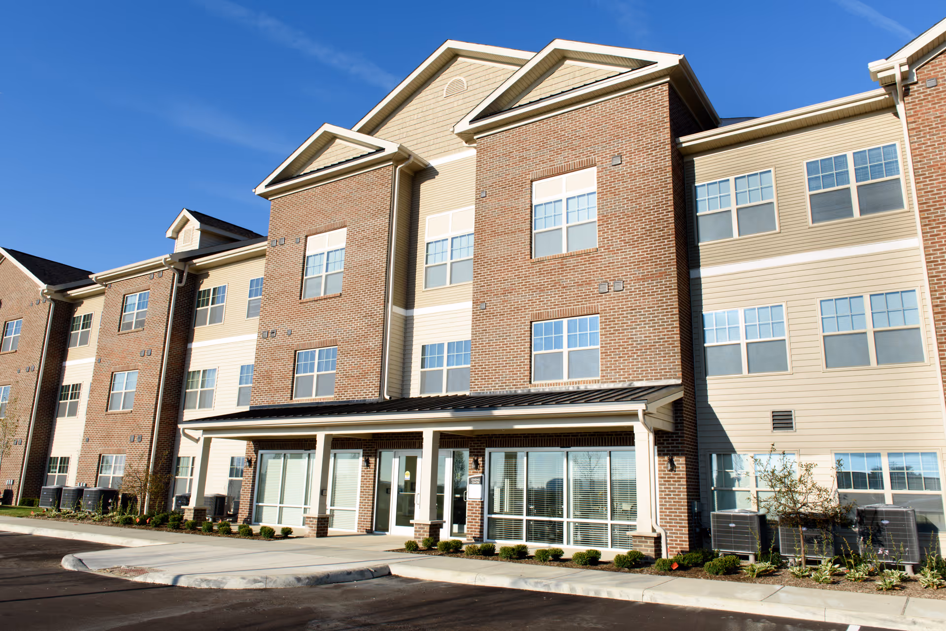 Exterior view of a three-story senior living facility building with brick and beige siding, multiple windows, and a covered entrance under a clear blue sky.