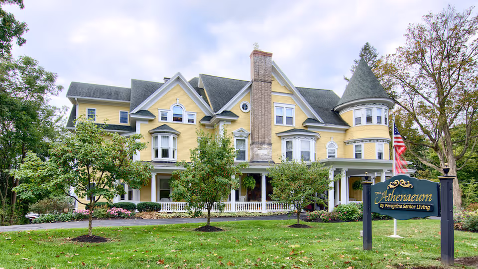 A large, yellow Victorian-style building with multiple gables, a turret, and a prominent chimney. The building is surrounded by green grass, trees, and shrubs. In front of the building is a sign that reads 'The Athenaeum by Peregrine Senior Living'. An American flag is visible near the sign.