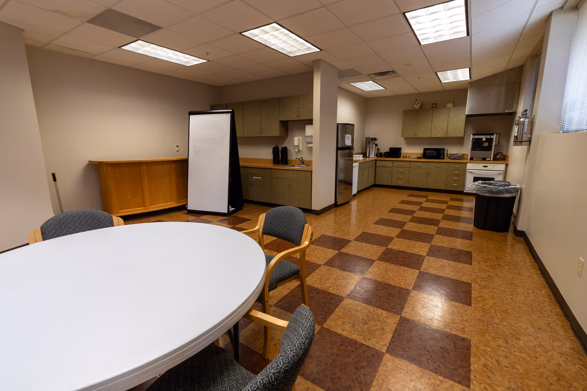 A spacious kitchen and dining area with a round white table surrounded by four chairs in the foreground. The kitchen has green cabinets, a stainless steel refrigerator, microwave, oven, and coffee maker. The floor has a checkered pattern with brown and tan tiles. The ceiling has fluorescent lighting, and there is a large trash bin near the window with blinds.