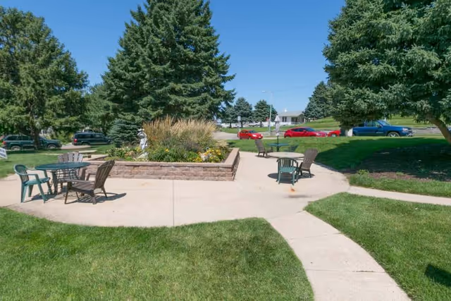 Sunlit outdoor courtyard with patio tables and chairs surrounding a raised flower bed and large evergreen trees.