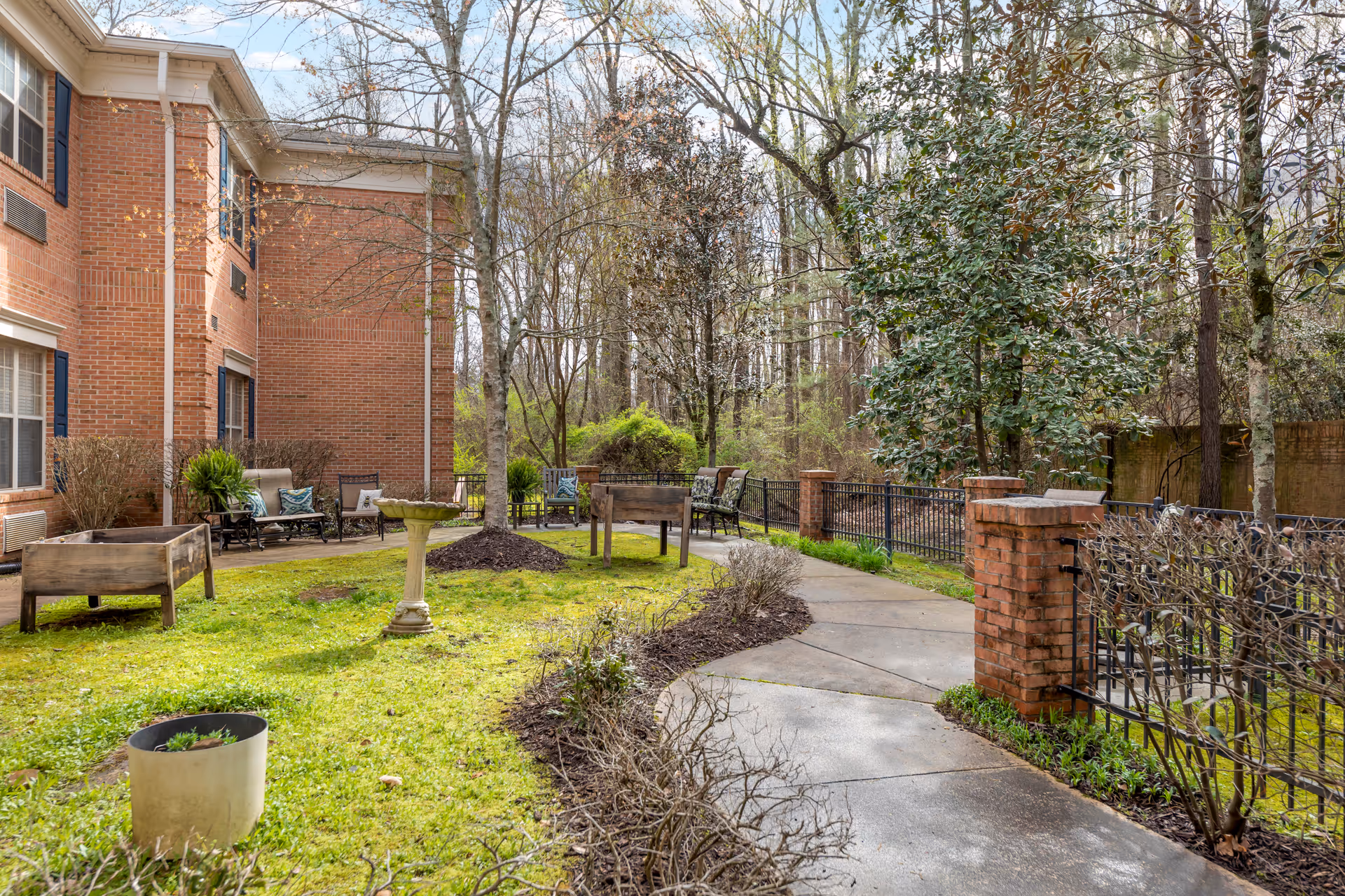 Outdoor garden area at Brookdale Germantown featuring a paved walkway, green grass, several trees, raised garden beds, a birdbath, and seating with cushioned chairs near a brick building.