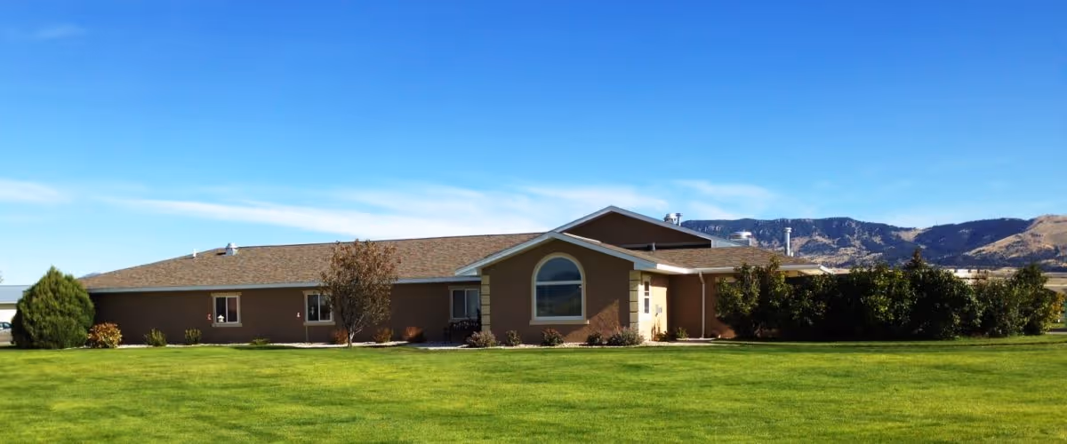 Single-story building with a brown exterior and a large arched window, surrounded by a well-maintained green lawn and some bushes and trees, with mountains visible in the background under a clear blue sky.