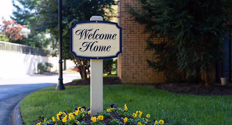 A decorative sign reading 'Welcome Home' is mounted on a post in a flower bed with yellow and purple flowers. The background shows a brick building, green grass, trees, and a paved driveway or walkway.
