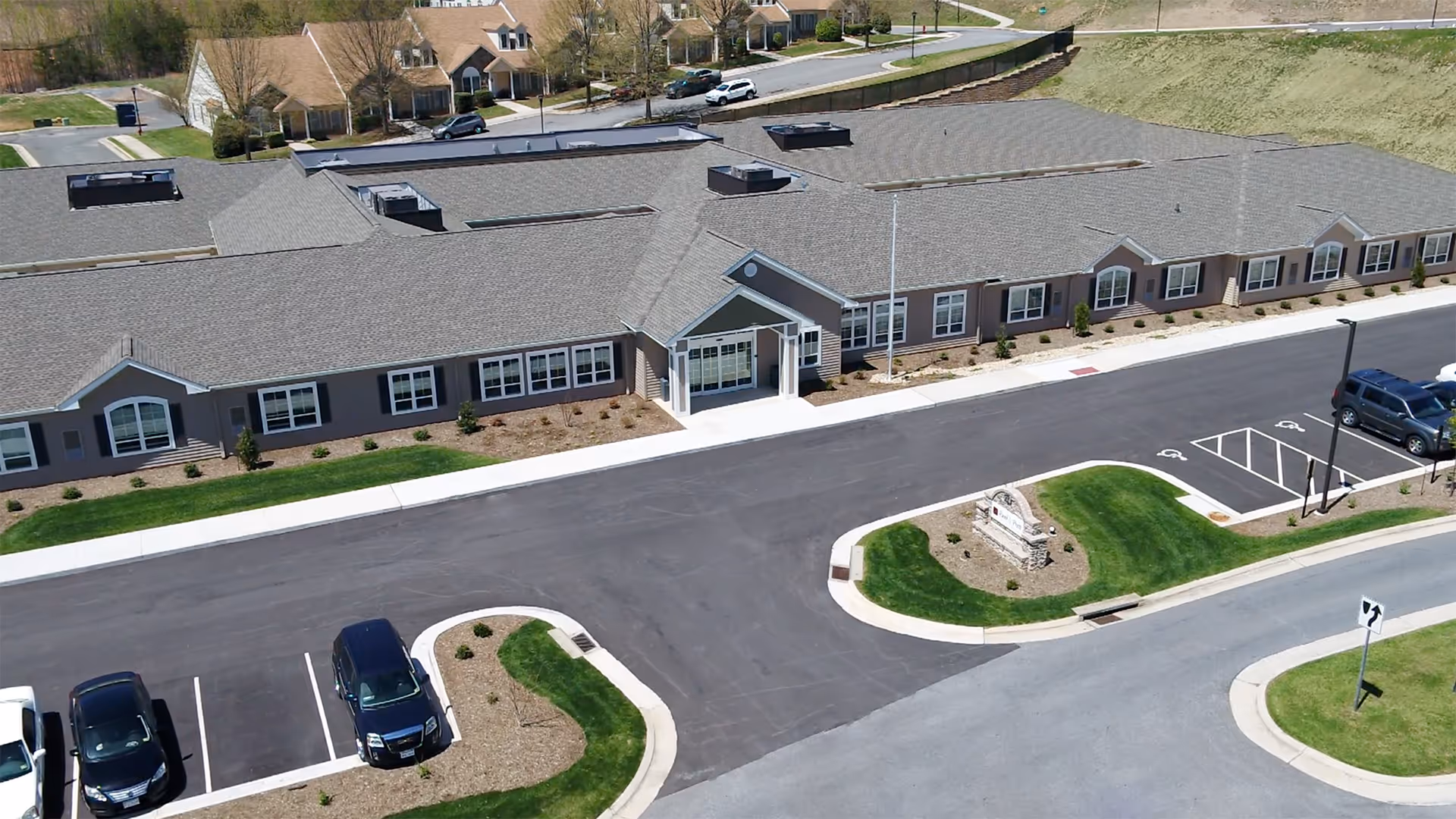 Aerial view of a single-story senior living facility building with a gray roof and multiple windows. The building has a main entrance with a small covered porch. There is a parking lot with several cars parked and landscaped areas with grass and small bushes surrounding the building. A sign is visible near the entrance on a grassy island.