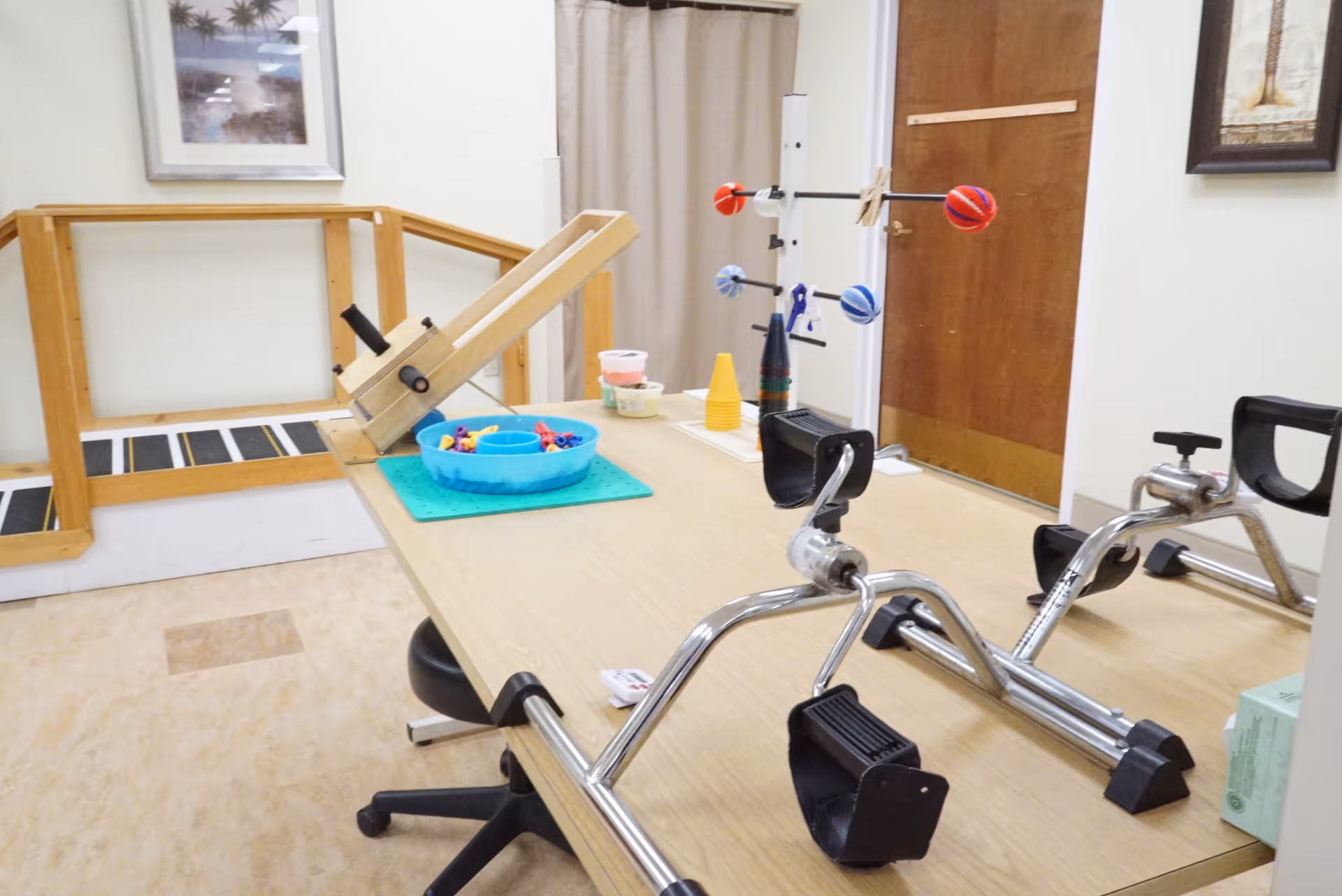 A therapy or rehabilitation room with a wooden table holding various exercise and therapy equipment including hand pedals, a ramp, colorful balls on a stand, and small containers. The room has a beige floor, light-colored walls, a wooden door, and framed pictures on the walls.