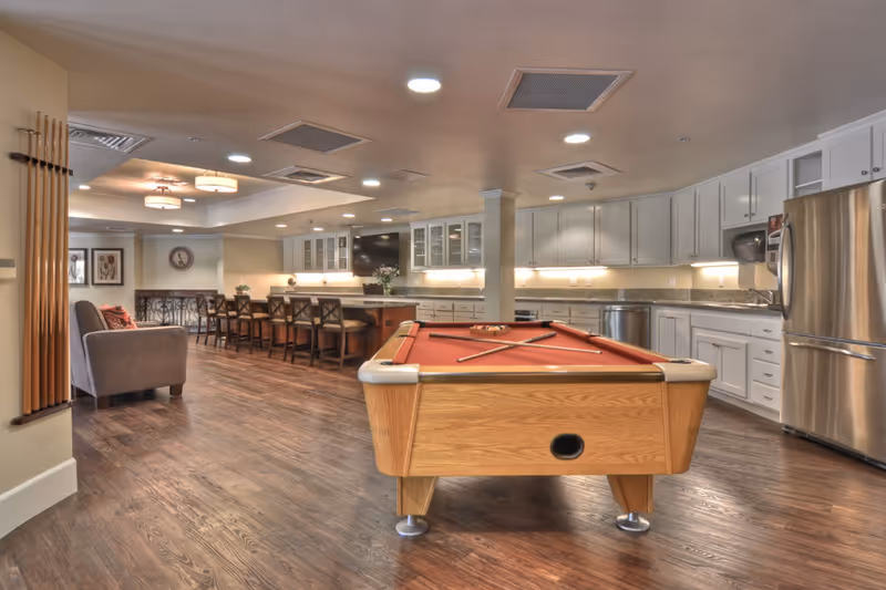 A spacious common area featuring a pool table with cues and balls, a long kitchen counter with bar stools, white cabinetry, stainless steel refrigerator, and a seating area with a gray armchair. The room has wood flooring and recessed lighting.