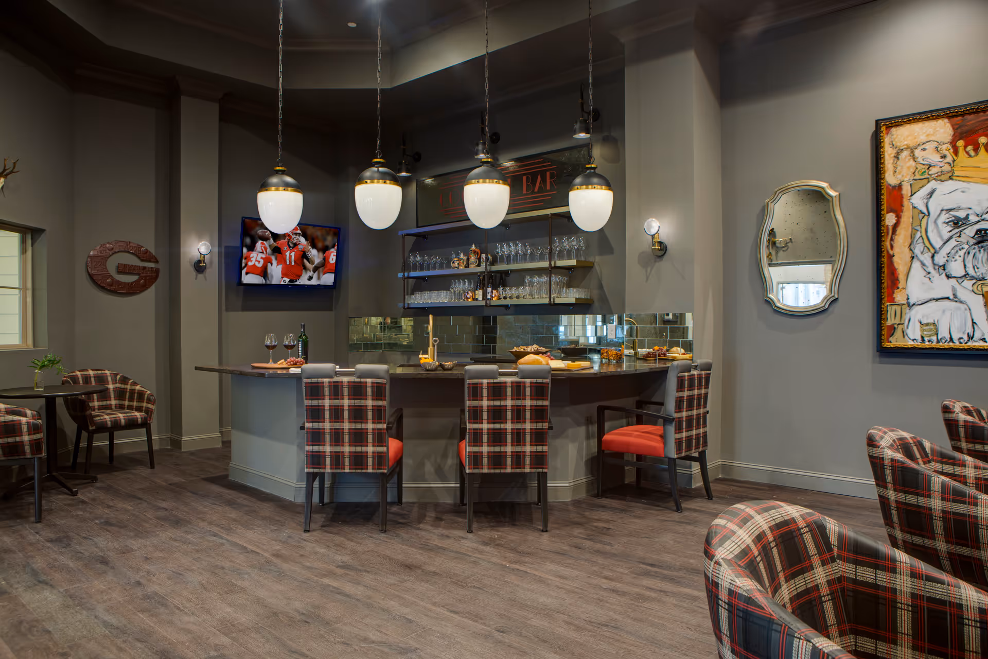 A cozy lounge area with a bar counter featuring four plaid chairs with red cushions. Above the bar, four pendant lights hang from the ceiling. Behind the bar, shelves hold various glasses and bottles. A TV mounted on the wall shows a football game. The room has gray walls, a wooden floor, a decorative mirror, and a colorful painting of a bulldog wearing a crown. Additional plaid chairs and small tables are visible around the room.