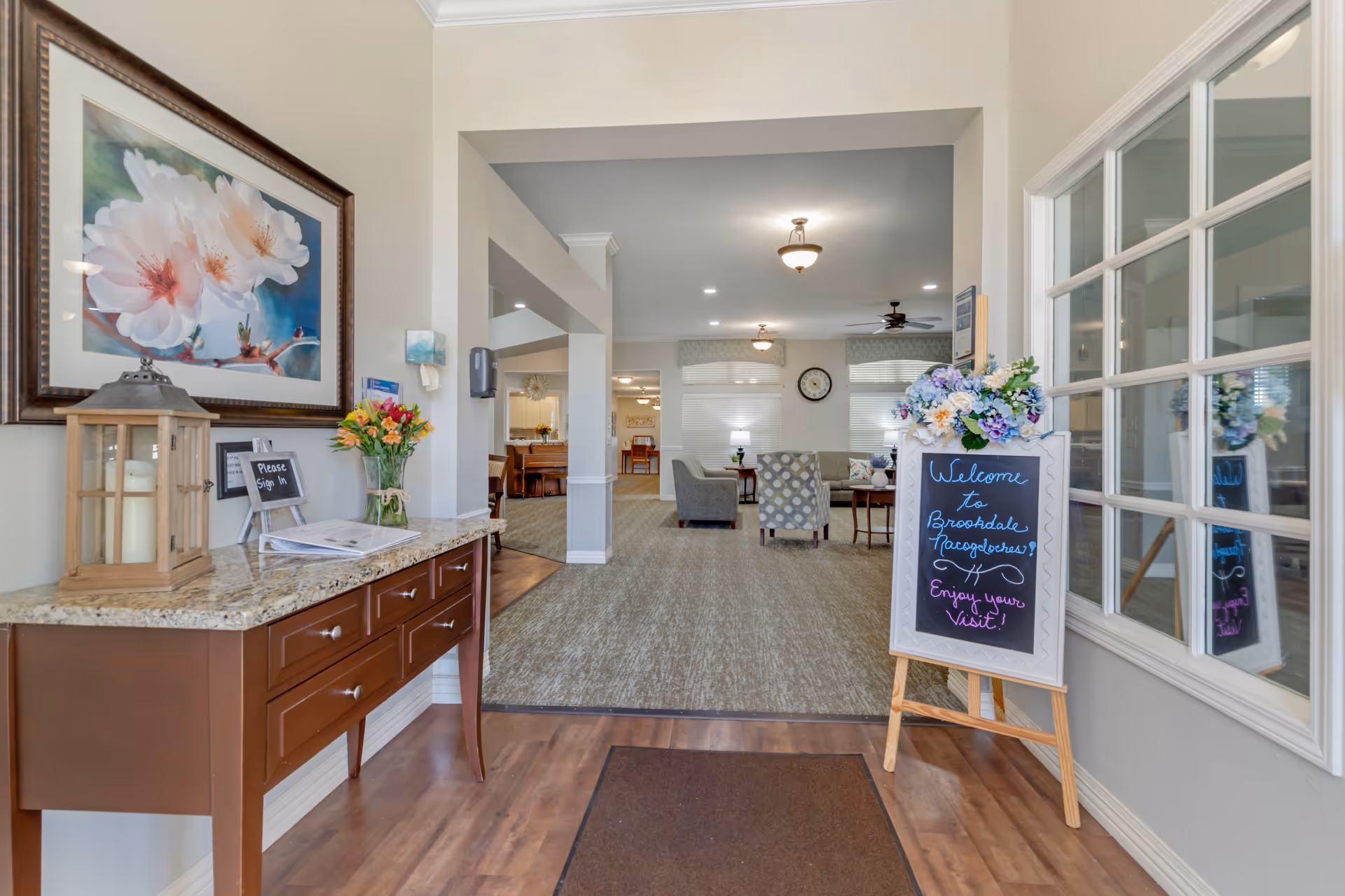 Entrance area of Brookdale Nacogdoches senior living facility featuring a wooden console table with a candle lantern, a vase of flowers, and a sign-in book. A chalkboard sign on an easel welcomes visitors. The view extends into a carpeted common area with seating, a clock on the wall, and soft lighting.