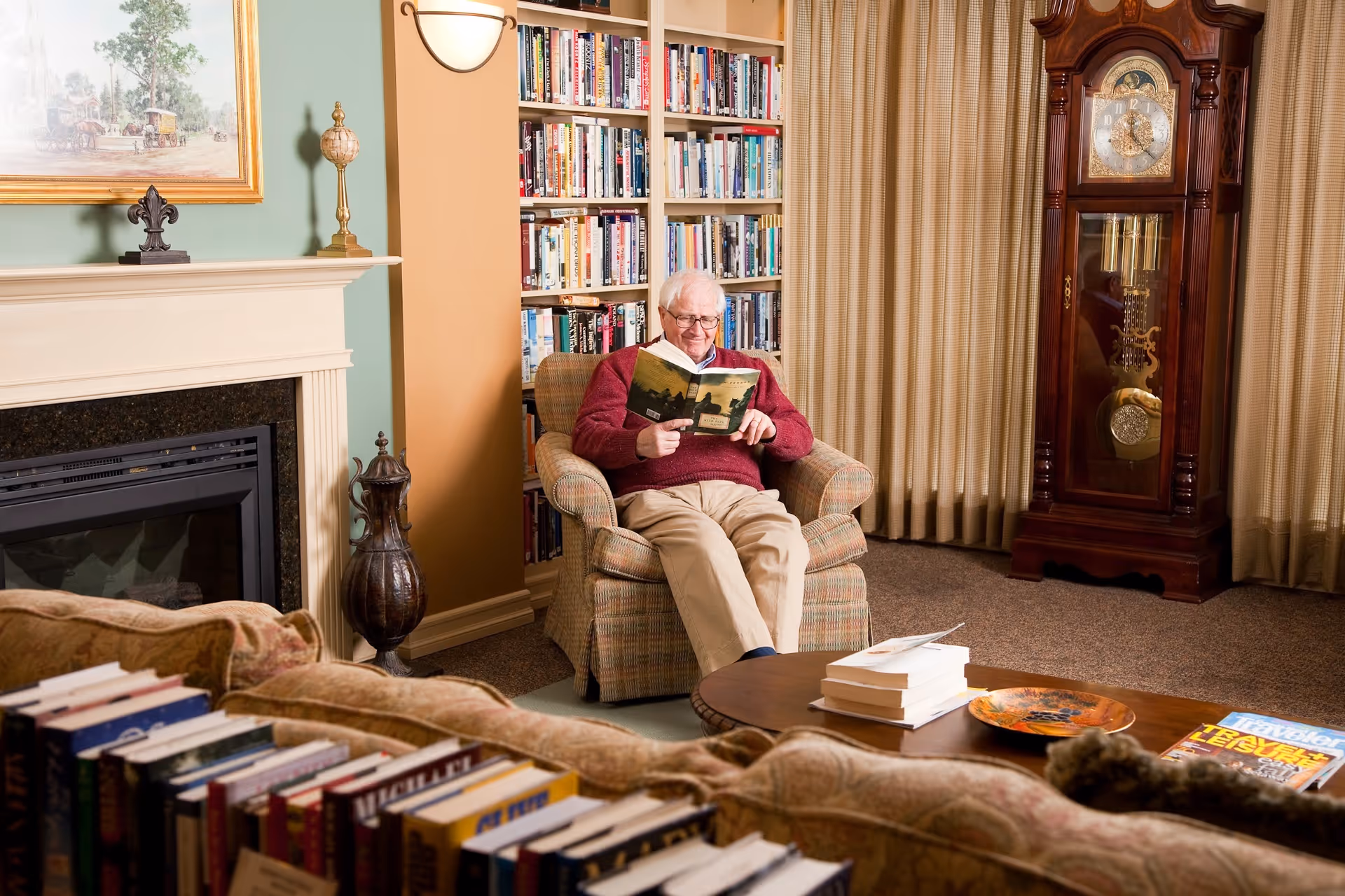 An elderly man wearing a red sweater and beige pants is sitting in a comfortable armchair reading a book in a cozy living room. The room features a fireplace with decorative items on the mantel, a large bookshelf filled with books, a wooden grandfather clock, and a round coffee table with books and a decorative plate. Beige curtains cover the windows behind the clock.