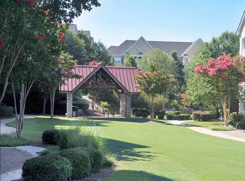 A landscaped courtyard with a red-roofed wooden pavilion, manicured lawn, walking paths, and apartment buildings in the background.