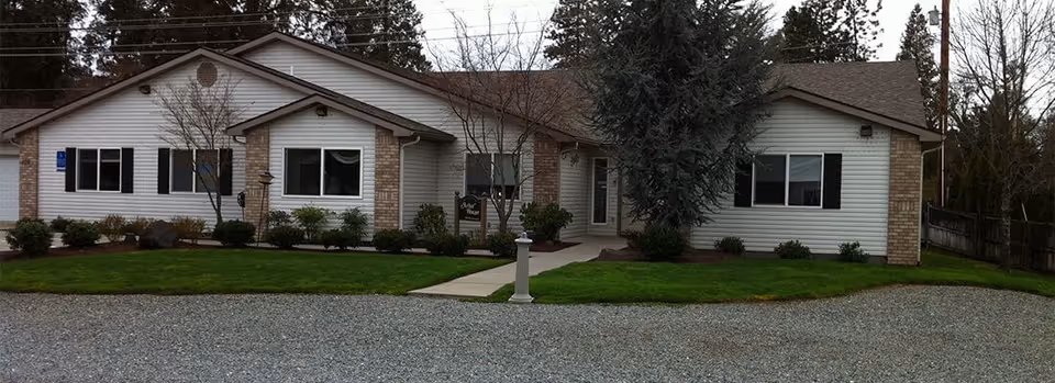 Single-story building with white siding and brick accents, surrounded by a well-maintained lawn and small shrubs, with a gravel driveway in front and trees in the background under an overcast sky.