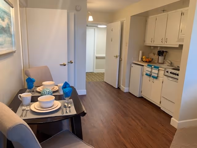 A small dining area with a table set for two, featuring beige chairs, white dishes, and blue napkins. To the right is a compact kitchen with white cabinets, a small refrigerator, stove, and countertop with utensils and a fruit bowl. The floor is wood, and there are open doors leading to other rooms in the background.