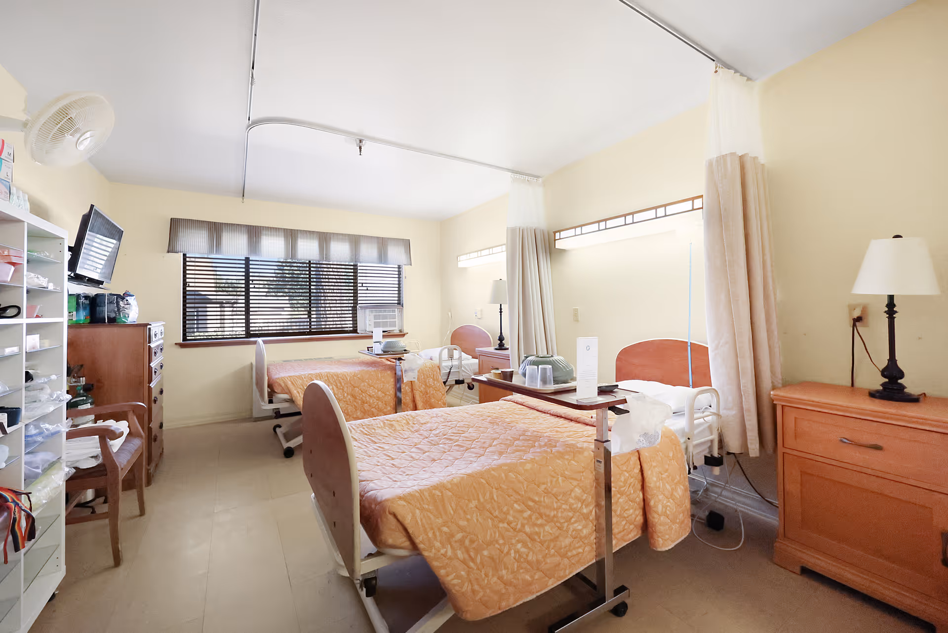 A bright room with two hospital-style beds covered in orange quilts, each with a bedside table and lamp. There is a window with blinds letting in natural light, a TV mounted on the wall, and shelves with various items on the left side. The room has light-colored walls and a clean, organized appearance.