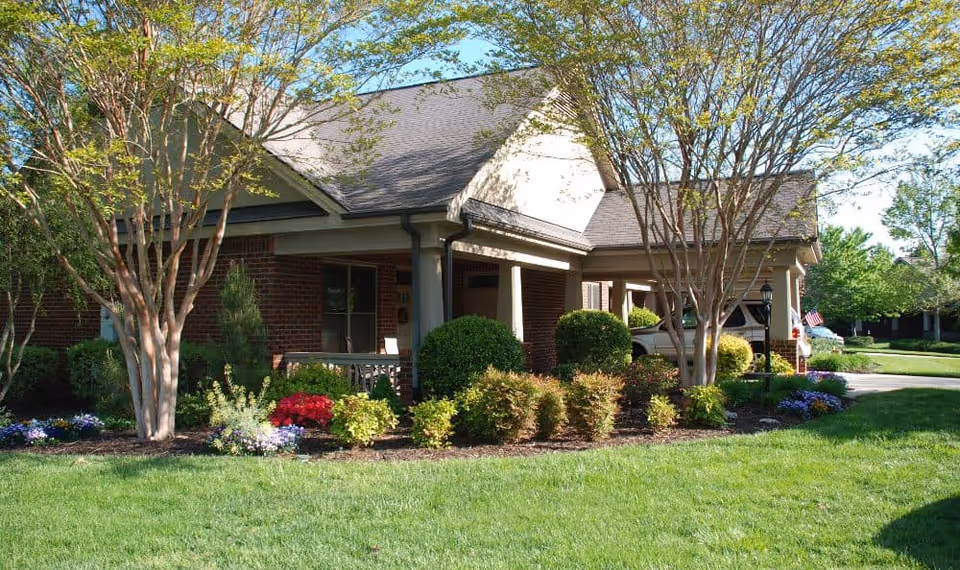 Exterior view of a single-story brick building with a covered porch, surrounded by well-maintained landscaping including bushes, flowering plants, and trees under a clear blue sky.