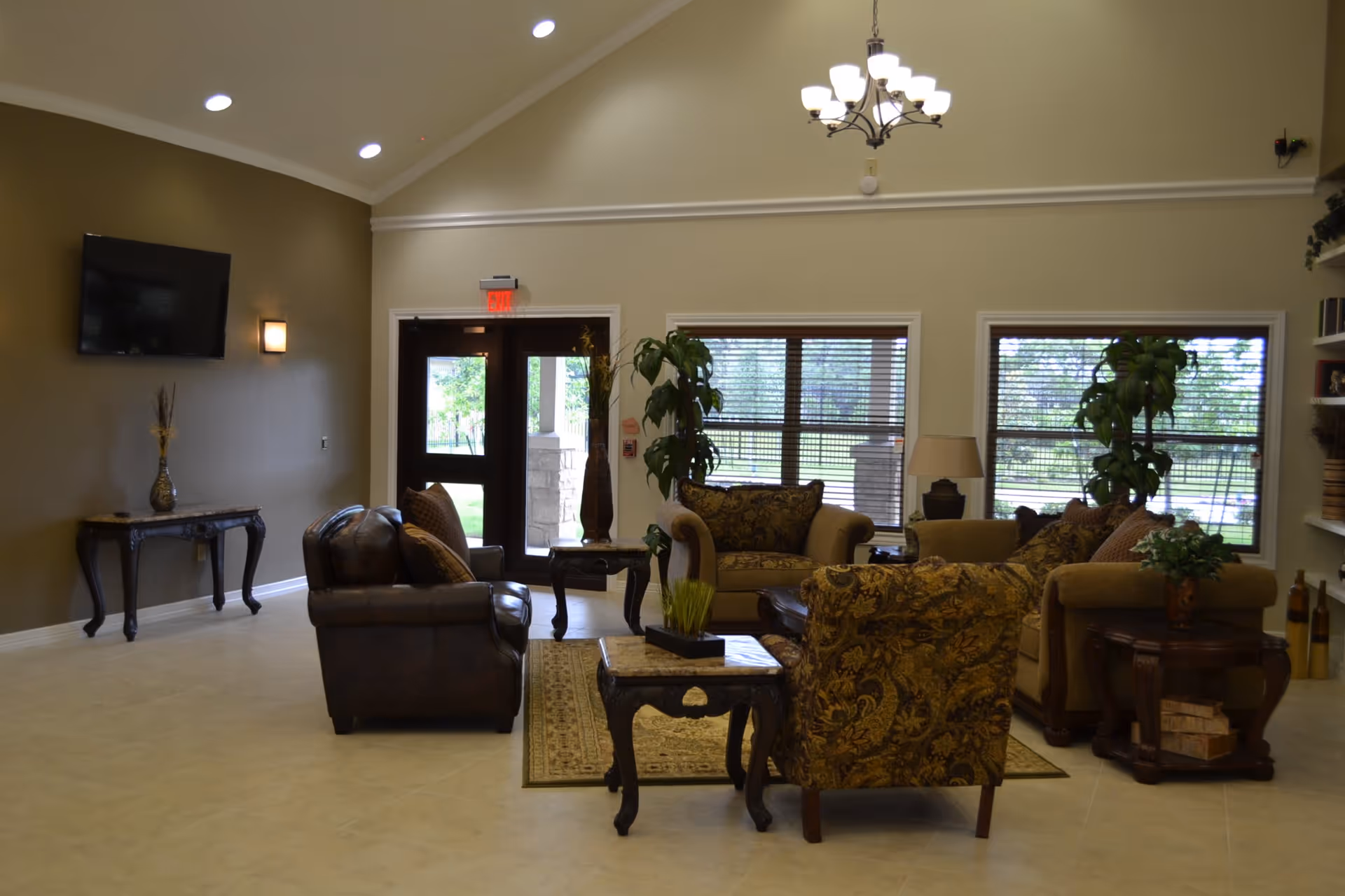 Bright communal living room with sofas and armchairs arranged around a coffee table, large windows, a chandelier and a wall-mounted TV.