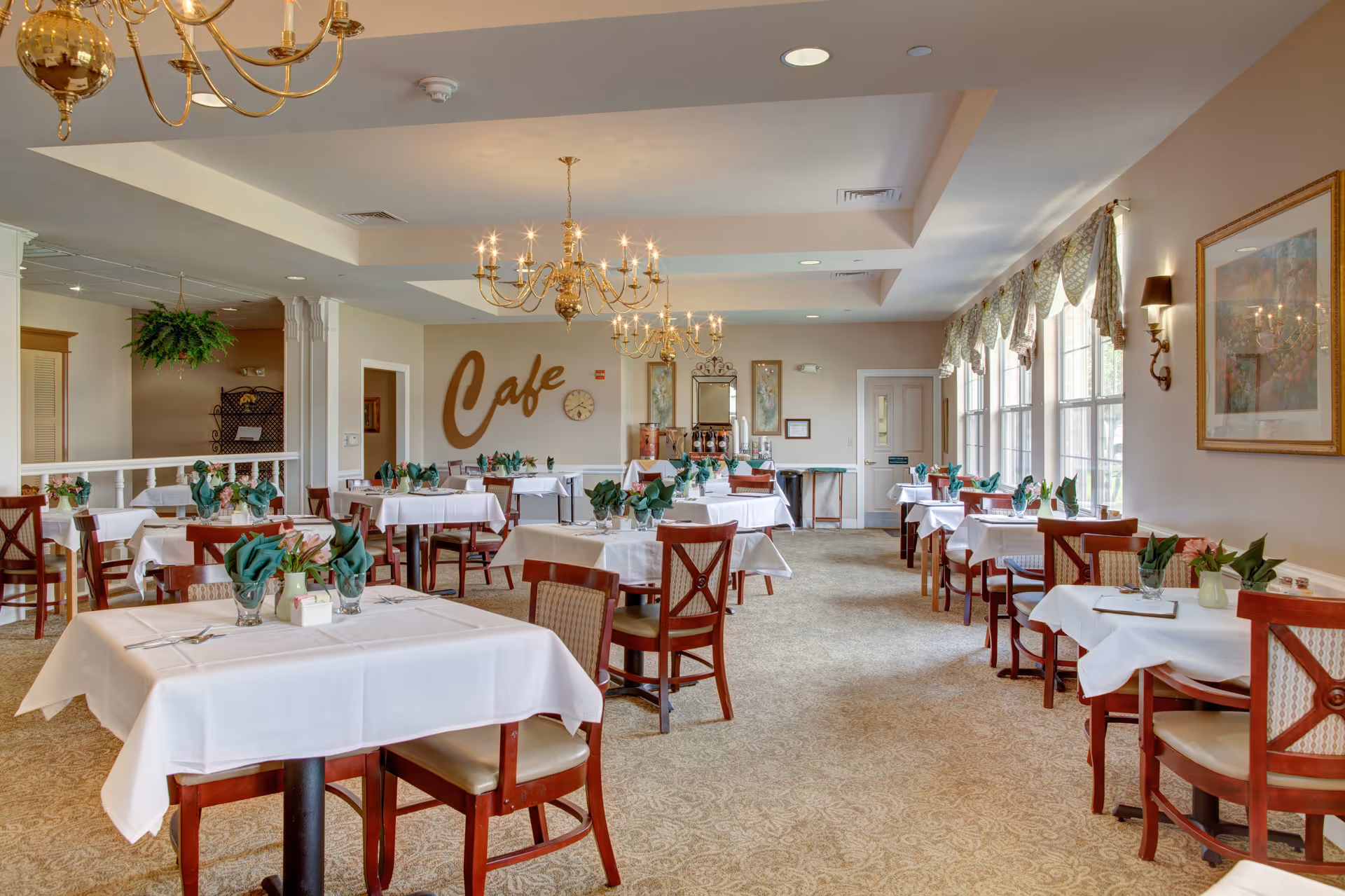 A bright and elegant dining room with multiple tables covered in white tablecloths and set with green folded napkins and small floral centerpieces. The room features large windows with patterned valances, beige carpet, and gold chandeliers hanging from a white ceiling. On the far wall, the word 'Cafe' is displayed in large decorative letters above a clock and beverage station.
