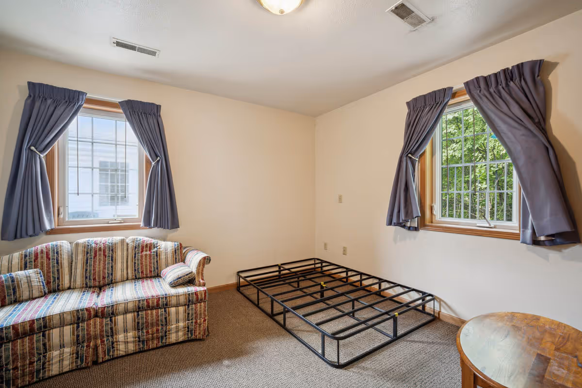 A small room with two windows covered by blue curtains. There is a striped couch on the left side, a metal bed frame without a mattress in the center, and a small round wooden table on the right. The walls are plain and light-colored, and the floor is carpeted.