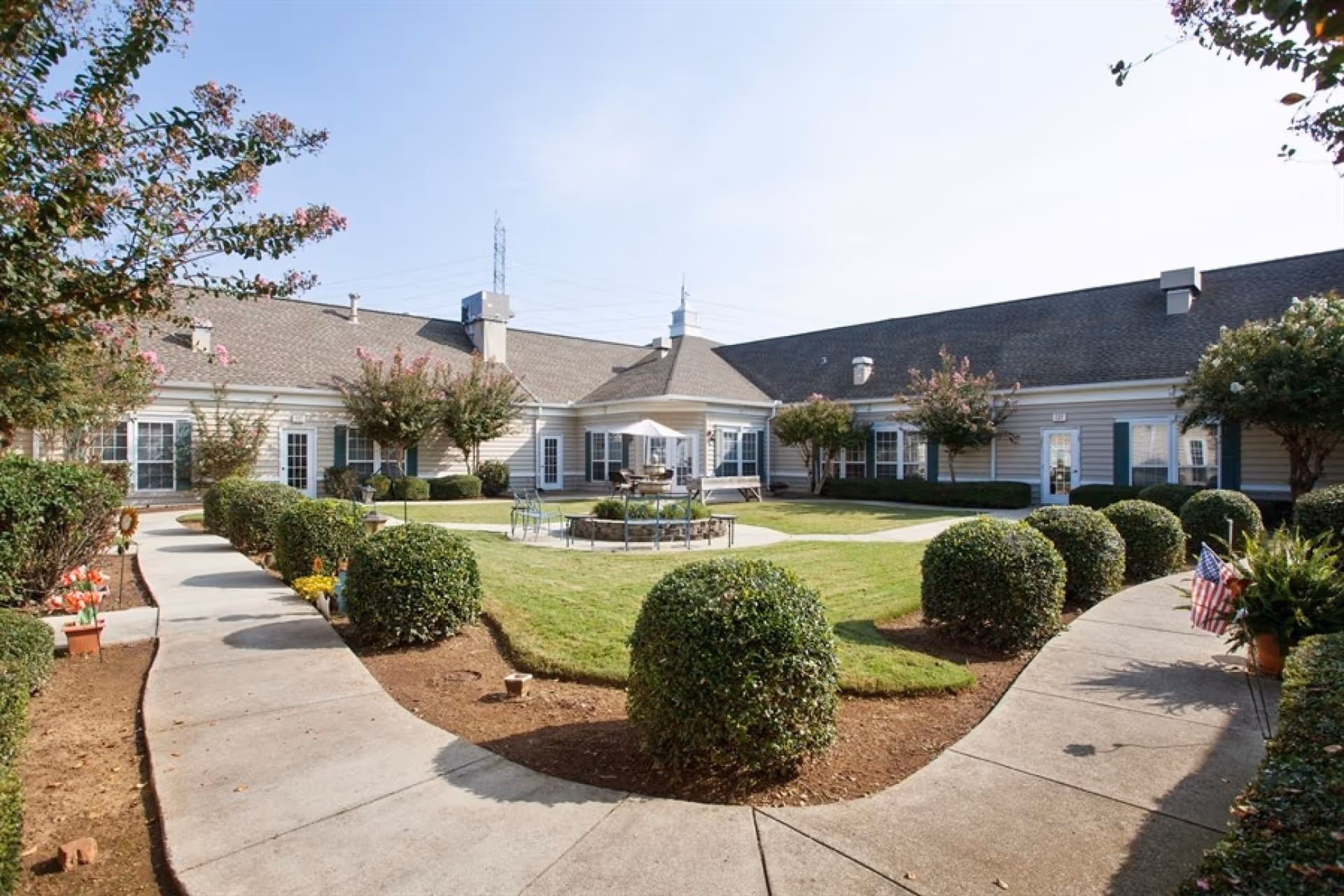 Outdoor courtyard area of a senior living facility with a circular walkway, neatly trimmed bushes, green lawn, and a central seating area with a table and umbrella. The building surrounds the courtyard with multiple windows and doors, and there are flowering trees and plants along the paths.
