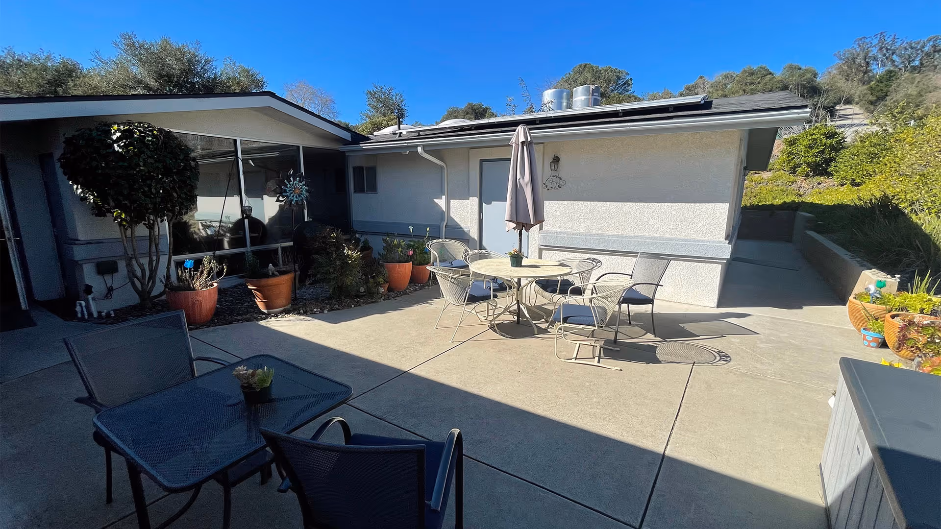 Outdoor patio area at Oak Park Manor Elderly Care Facility with two sets of tables and chairs. One table has an umbrella, and there are several potted plants around the patio. The building exterior is visible with a clear blue sky above.
