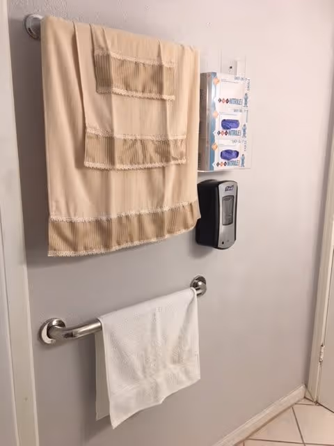 A wall with two towel racks holding beige and white towels, a wall-mounted hand sanitizer dispenser, and a box of disposable gloves in a clear holder.