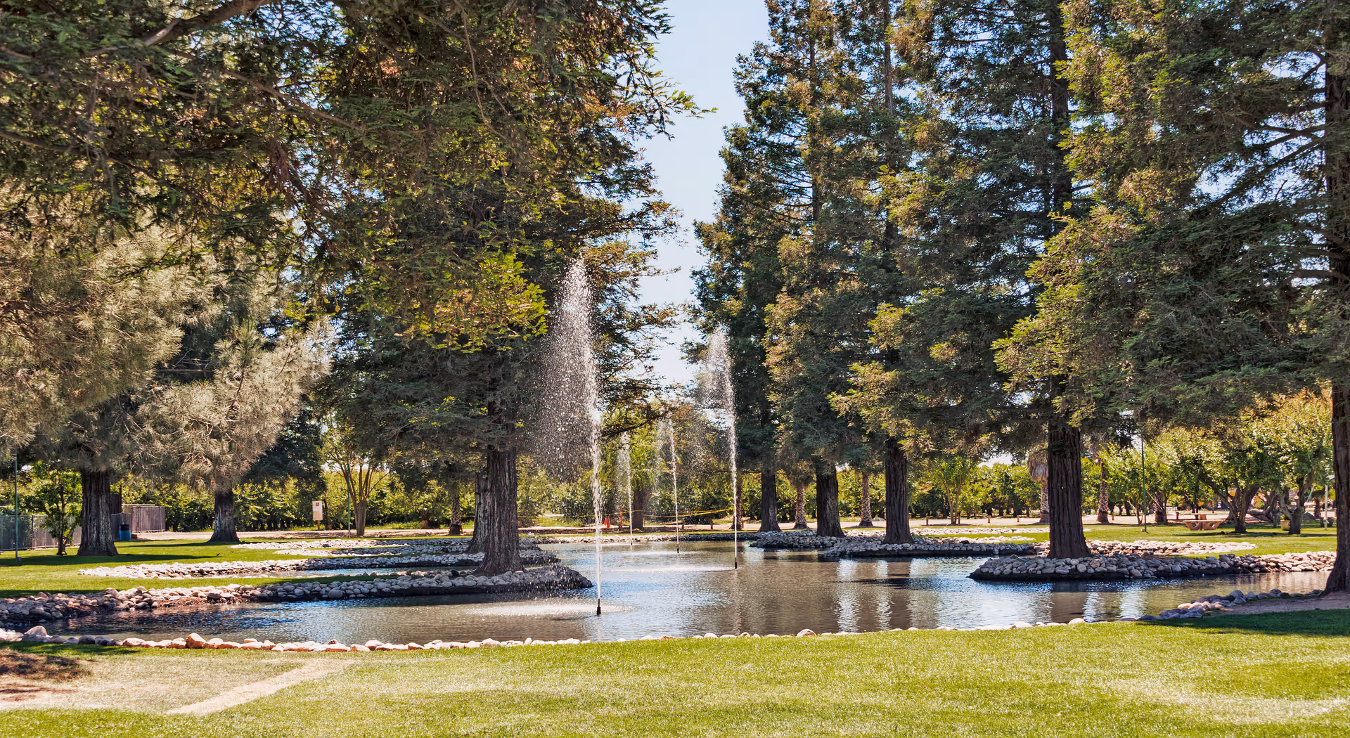 A landscaped outdoor pond with fountains surrounded by large trees and grassy lawns.