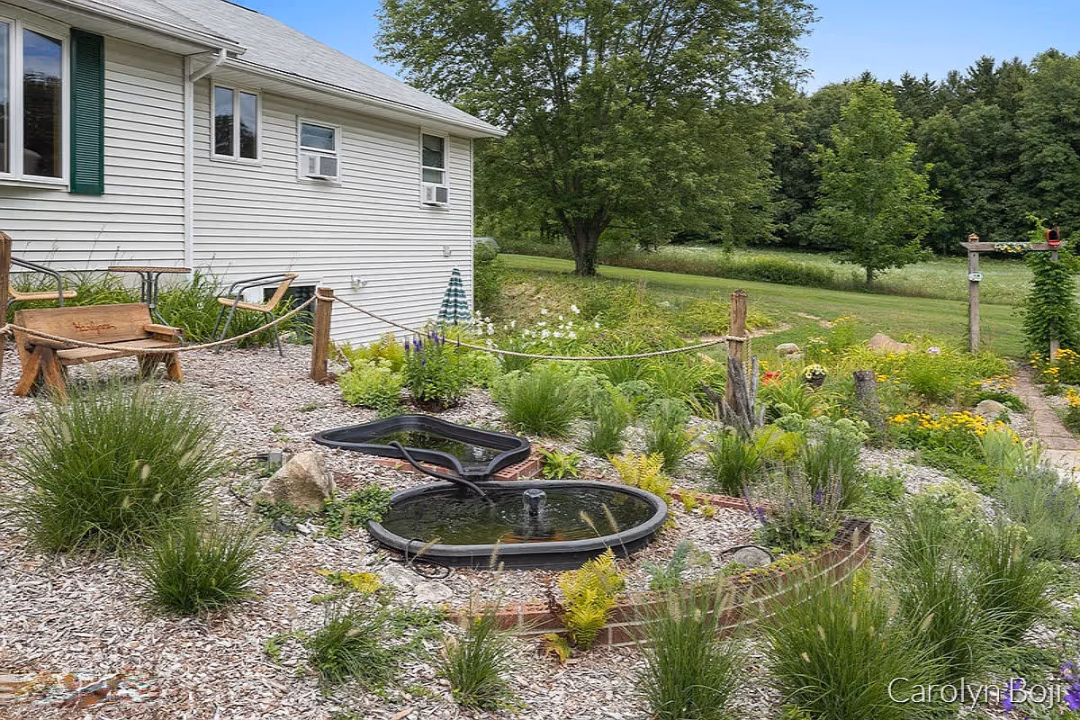 A landscaped garden area next to a white building with green shutters. The garden features a small pond with a fountain, various plants and flowers, a wooden bench, and a rope fence. Trees and a grassy field are visible in the background.