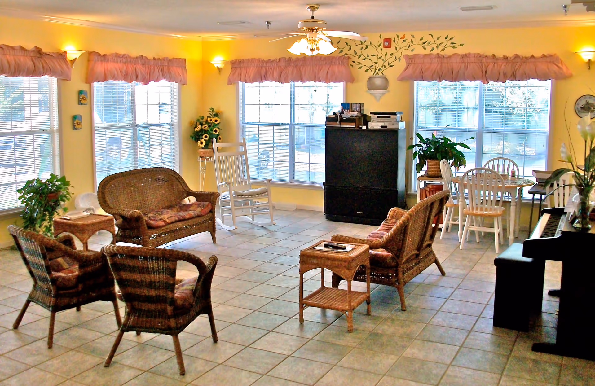 A cozy living room area with wicker chairs and a loveseat arranged around a small wicker table. There is a white rocking chair near large windows with pink valances. The room has yellow walls with decorative wall sconces and a ceiling fan with lights. A black TV cabinet with electronic devices is positioned against the wall. A small dining table with white chairs and a potted plant is visible near the windows. A piano with a bench and a vase of flowers is on the right side of the room. The floor is tiled.