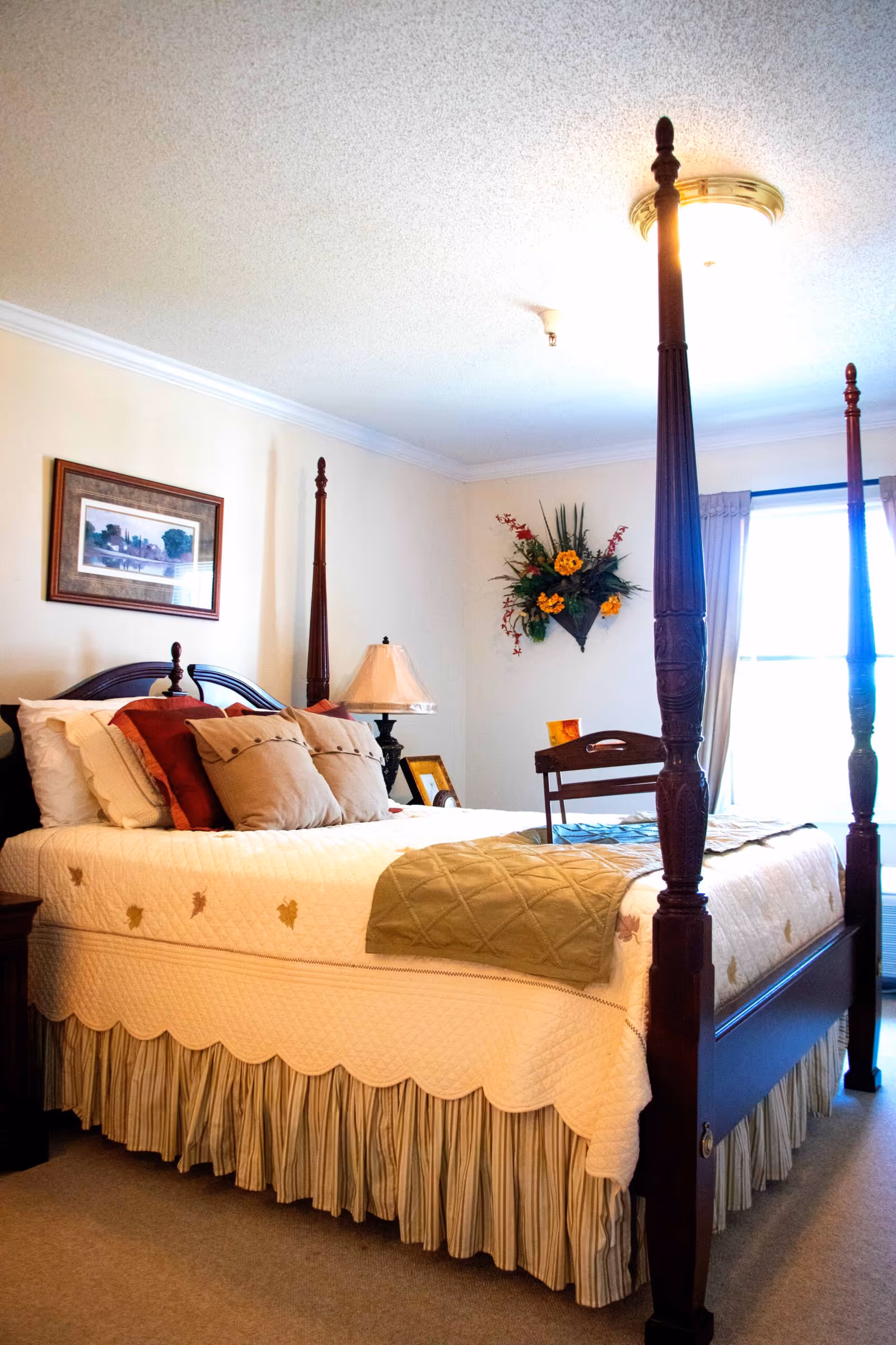 A cozy bedroom with a wooden four-poster bed dressed in a cream quilt and multiple pillows in beige and rust colors. A bedside table with a lamp and framed pictures is visible, along with a wall-mounted floral arrangement and a window with curtains letting in natural light.