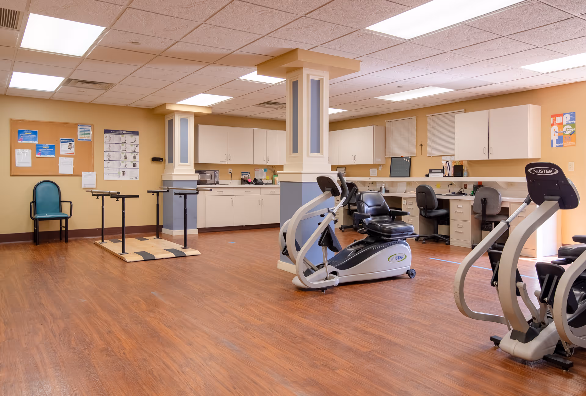 A spacious room with wooden flooring featuring exercise equipment including a recumbent bike and a NuStep machine. The room has beige walls with white cabinets and countertops along the back wall, and two workstations with chairs. There are two columns in the center of the room, a bulletin board with notices on the left wall, and a single chair against the wall. The ceiling has fluorescent lighting panels.