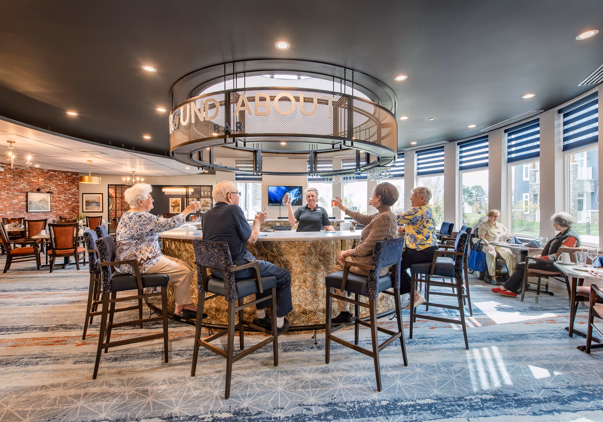 A group of elderly people sitting on high chairs around a circular bar counter inside a bright, modern lounge area with large windows and blue blinds. A bartender is serving drinks and the atmosphere is social and welcoming.