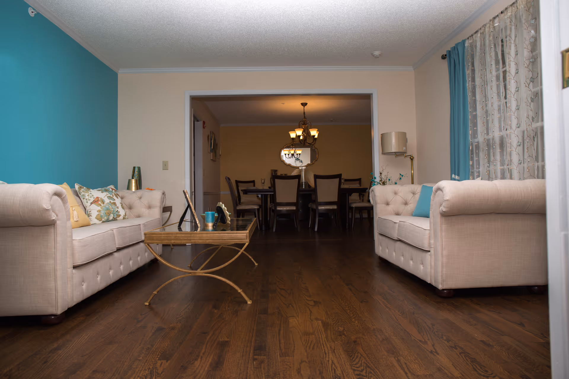 A cozy living room with two beige tufted sofas facing each other, a glass coffee table with a gold frame in the center, and a teal accent wall on the left. In the background, there is a dining area with a wooden dining table and chairs, a chandelier, and a decorative mirror on the wall. The room has wooden flooring and a window with sheer curtains and teal drapes on the right.