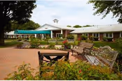 Outdoor patio area of Avon Health Center featuring several benches and chairs arranged on a brick-paved surface, surrounded by greenery and trees, with the single-story facility building visible in the background under a partly cloudy sky.