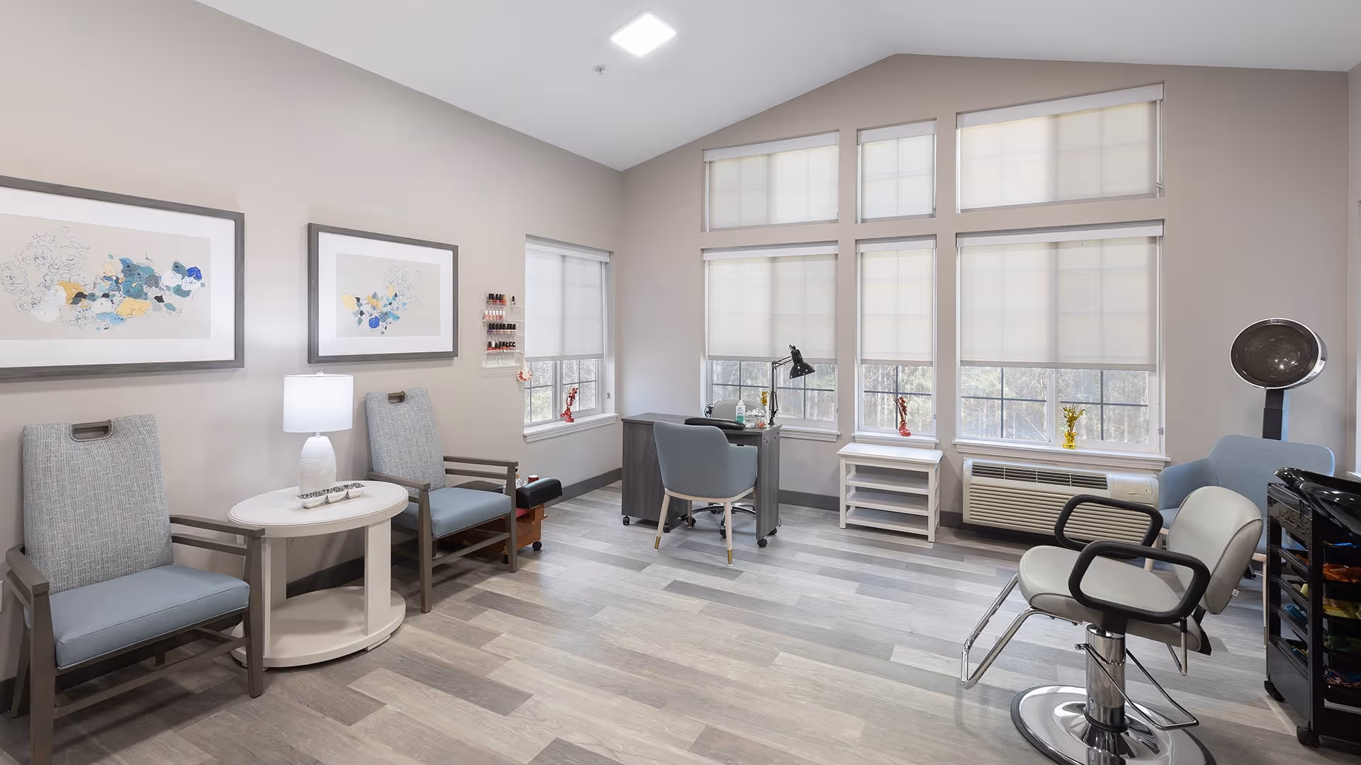 A bright and clean salon room with two framed abstract artworks on the wall, two cushioned chairs with armrests beside a small round table with a lamp, a desk with a chair near large windows with blinds, a salon hair dryer chair, and a rolling cart with supplies.