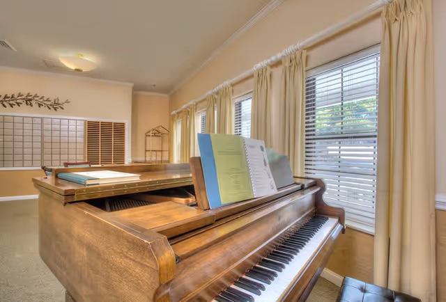Interior room with a wooden grand piano in the foreground, sheet music on the piano, beige walls, multiple windows with cream-colored curtains, and a wall of mailboxes in the background.