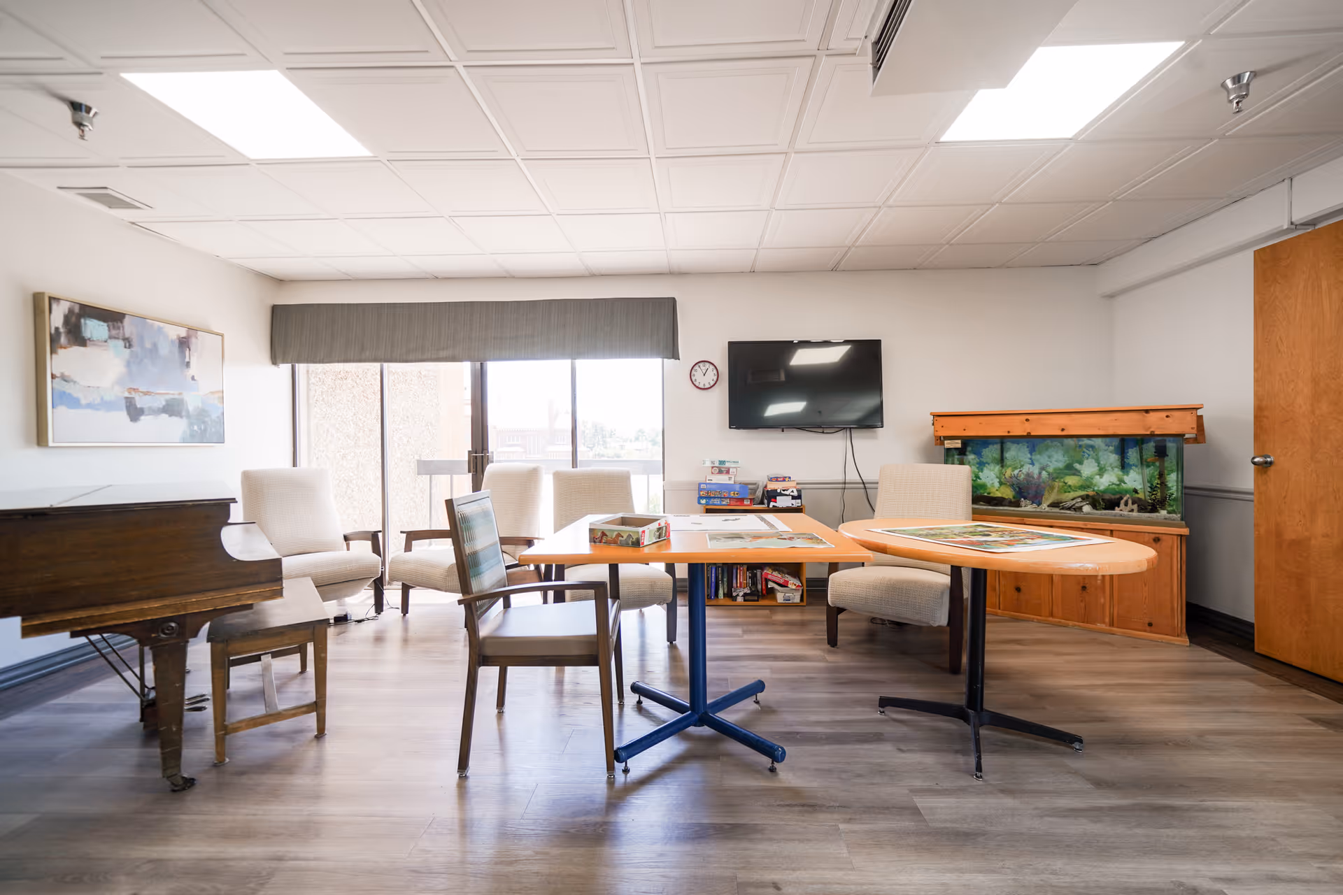 Bright communal activity room with chairs and tables, a piano, a fish tank, and a wall-mounted TV.