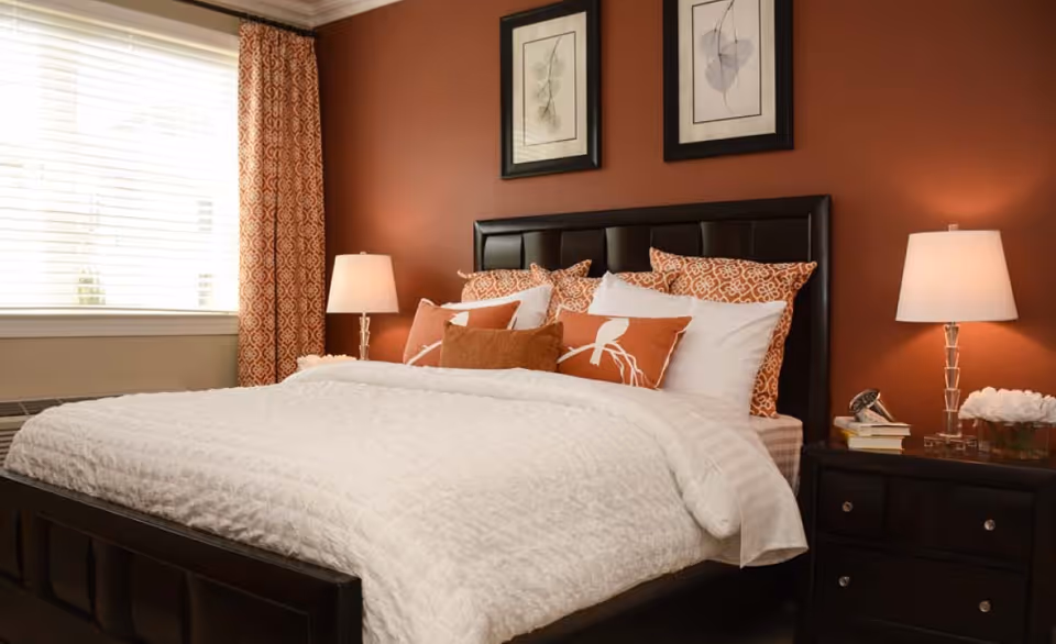 A cozy bedroom with a large bed featuring a dark leather headboard and footboard. The bed is made with white bedding and decorated with multiple orange and white patterned pillows. On either side of the bed are dark wooden nightstands with table lamps. The wall behind the bed is painted a warm orange color and has two framed botanical prints. A window with white blinds and orange patterned curtains is visible on the left side.