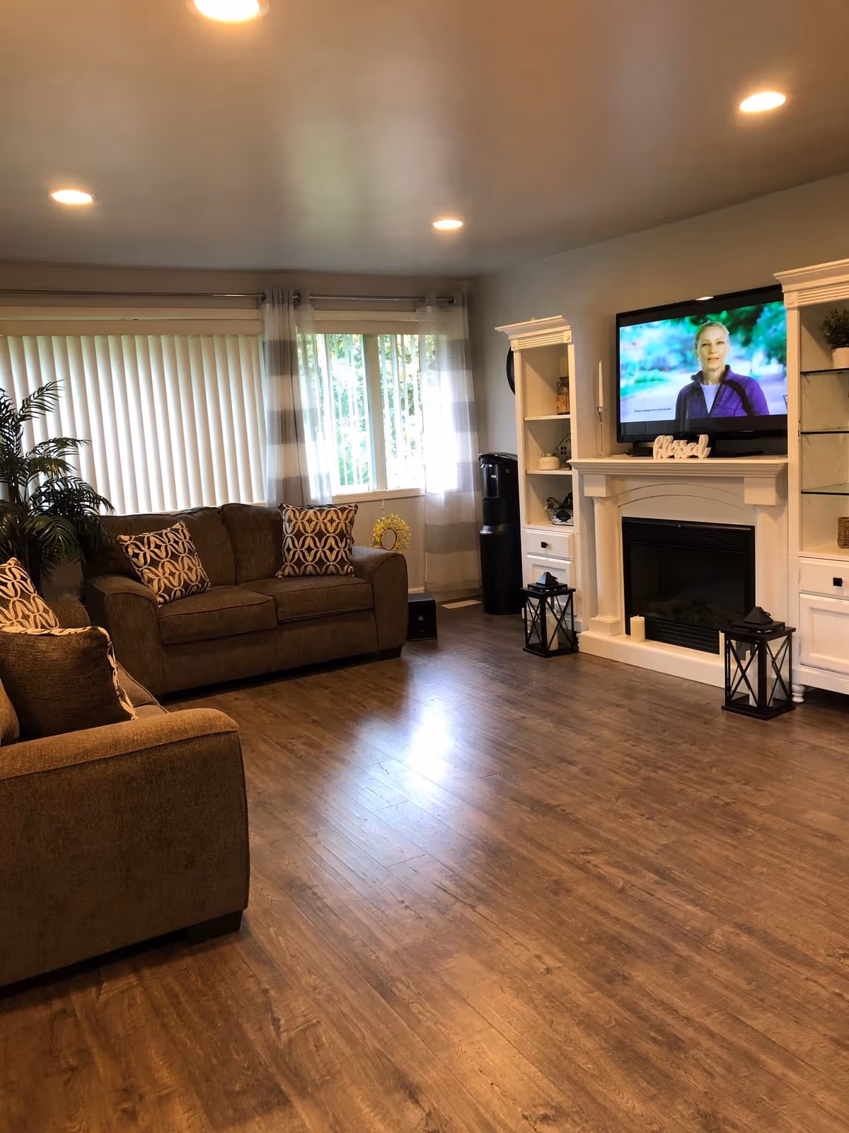 A cozy living room with brown sofas adorned with patterned cushions, a large window with vertical blinds and sheer curtains, a white fireplace with a TV mounted above it displaying a woman, built-in white shelving units on either side of the fireplace, a water cooler in the corner, and wooden flooring.