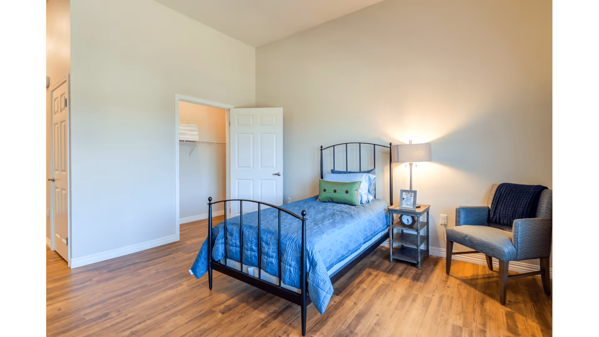 A bedroom with a single black metal bed frame with a blue quilt and pillows. Next to the bed is a small wooden nightstand with a lamp, a clock, and a framed photo. There is a gray upholstered chair with a dark blue throw blanket on it. The room has wooden flooring and light-colored walls. An open door reveals a walk-in closet with shelving.