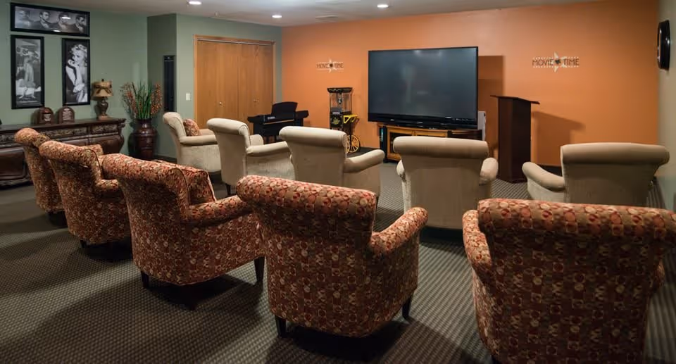 A cozy movie room with two rows of comfortable armchairs facing a large flat-screen TV mounted on an orange wall. The room has a carpeted floor, a popcorn machine, a small podium, and framed black-and-white photos on the green wall to the left.