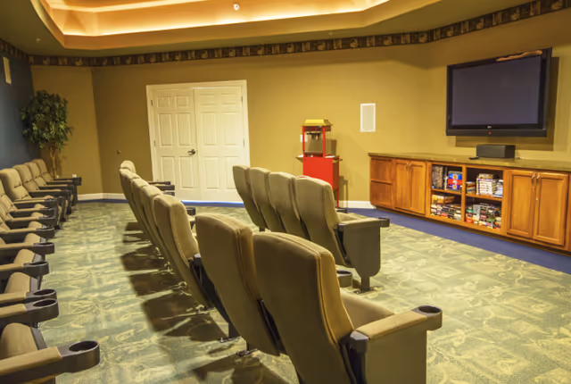 A small theater room with two rows of cushioned seats facing a wall-mounted flat screen TV. Below the TV is a wooden cabinet with shelves holding board games and DVDs. A red popcorn machine is placed against the back wall near double doors. The room has patterned carpet and warm lighting from a recessed ceiling.