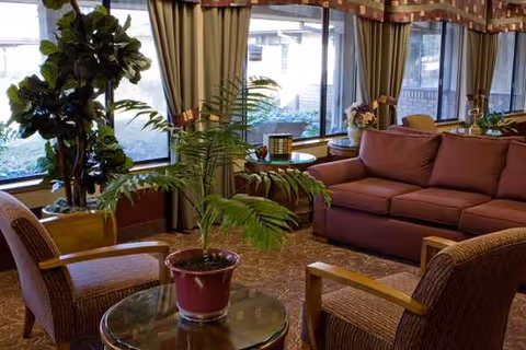 Cozy senior living lounge with a pink sectional sofa, chairs, plants, and large windows.
