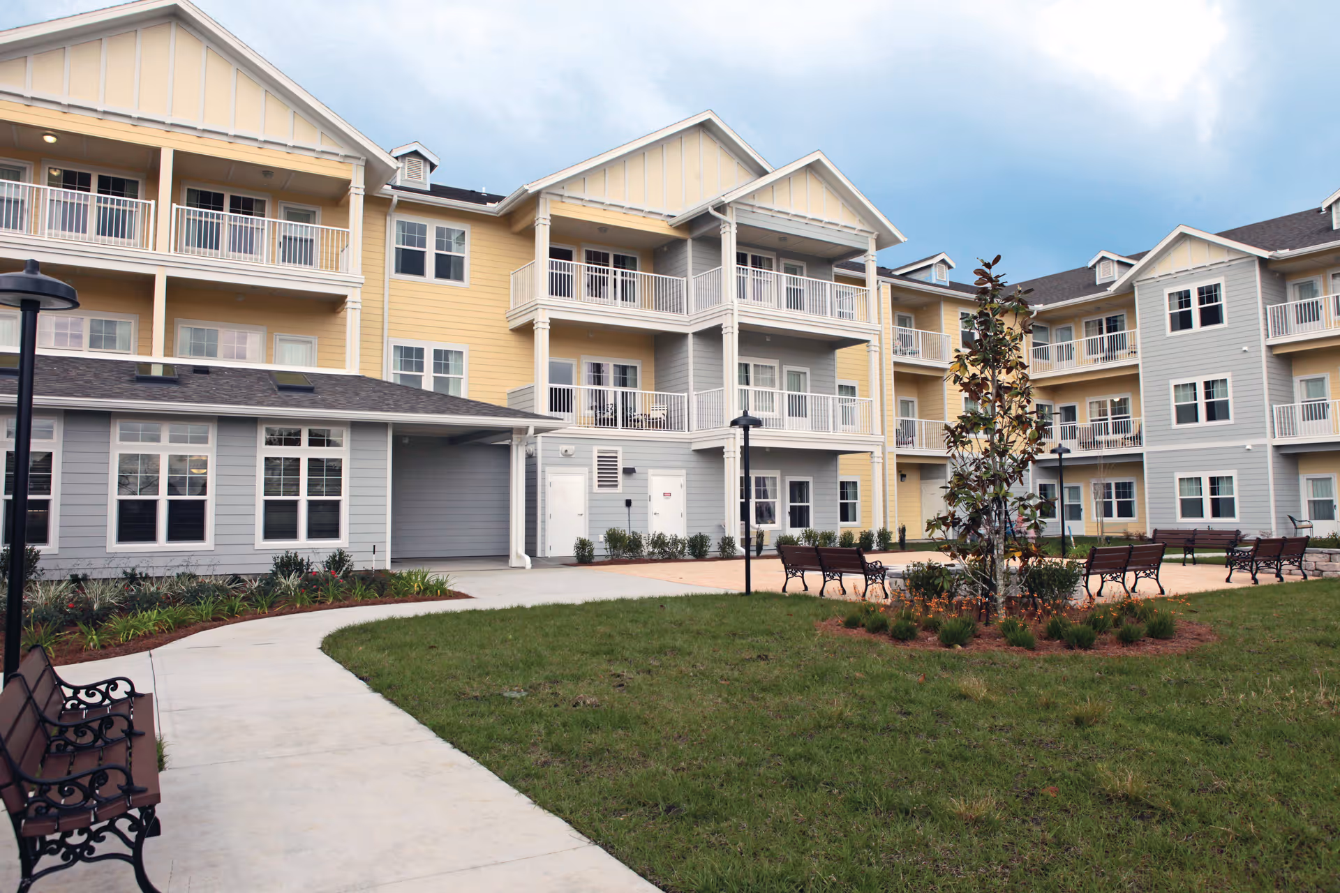 Exterior view of a multi-story senior living facility with yellow and gray siding, balconies, benches, a paved walkway, and landscaped greenery under a cloudy sky.