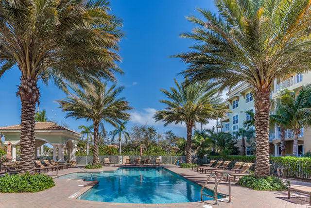 Outdoor swimming pool surrounded by lounge chairs, palm trees, a gazebo, and an adjacent multi-story building under a clear blue sky.