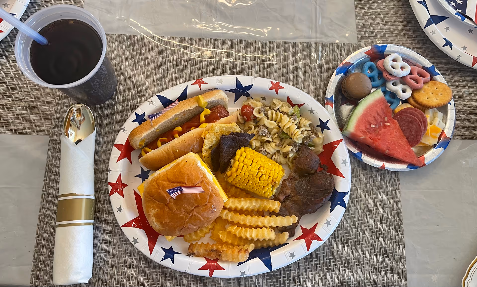 A patriotic-themed paper plate filled with a hot dog topped with mustard, a cheeseburger with a small American flag toothpick, crinkle-cut fries, corn on the cob, pasta salad, and grilled meat. Next to it is a smaller plate with a slice of watermelon, pretzels coated in red, white, and blue yogurt, crackers, salami, cheese cubes, and a chocolate truffle. A plastic cup with a dark beverage and a straw, along with a wrapped spoon and napkin, are placed on a woven placemat.