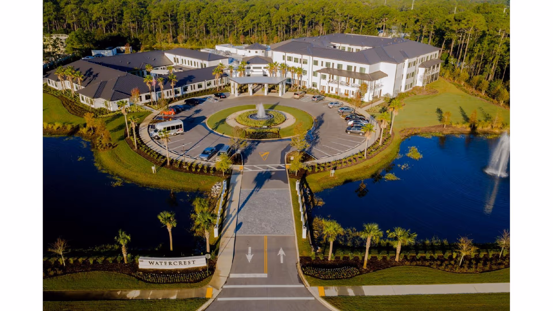 Aerial view of Watercrest Santa Rosa Beach senior living facility showing a large, modern building with a circular driveway and fountain at the entrance, surrounded by landscaped greenery, palm trees, and two water features with fountains on either side of the driveway.
