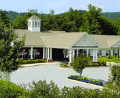 Exterior view of a single-story senior living facility building with a light-colored roof and white columns at the entrance. The building is surrounded by greenery, including trees and shrubs, with a circular driveway and landscaped flower beds in front.
