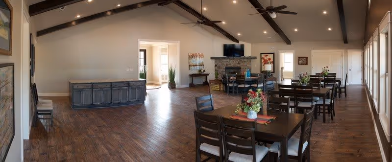 Spacious communal dining room with multiple dark wood tables and chairs, hardwood floors, exposed ceiling beams, and a stone fireplace with TV.