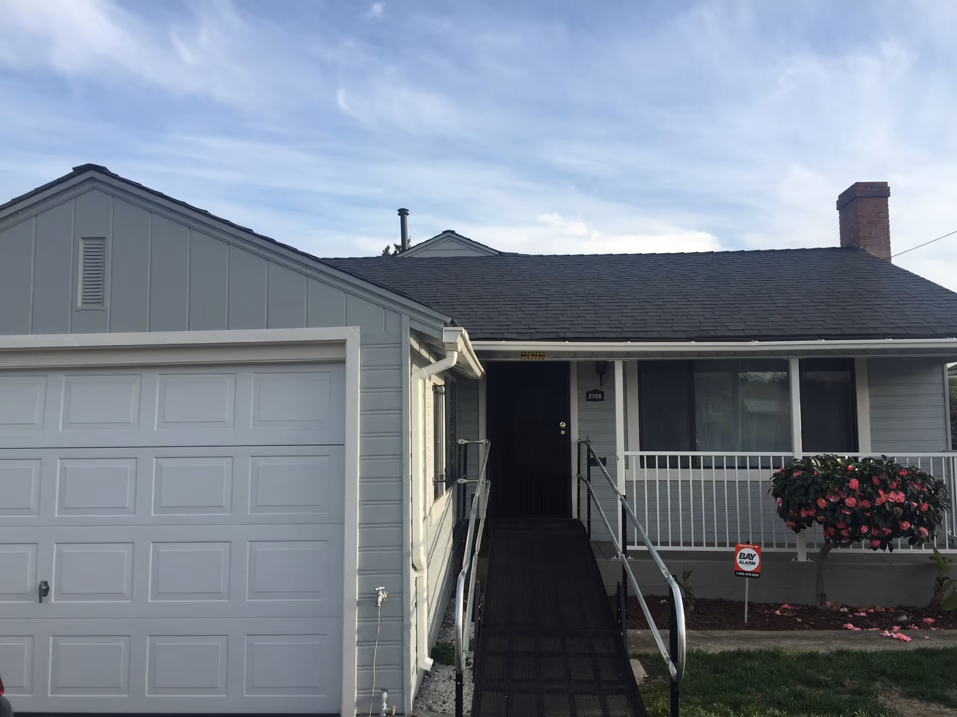Front exterior of a single-story light-gray house with an attached garage, a wheelchair ramp leading to the covered porch, and a flowering shrub by the entrance.