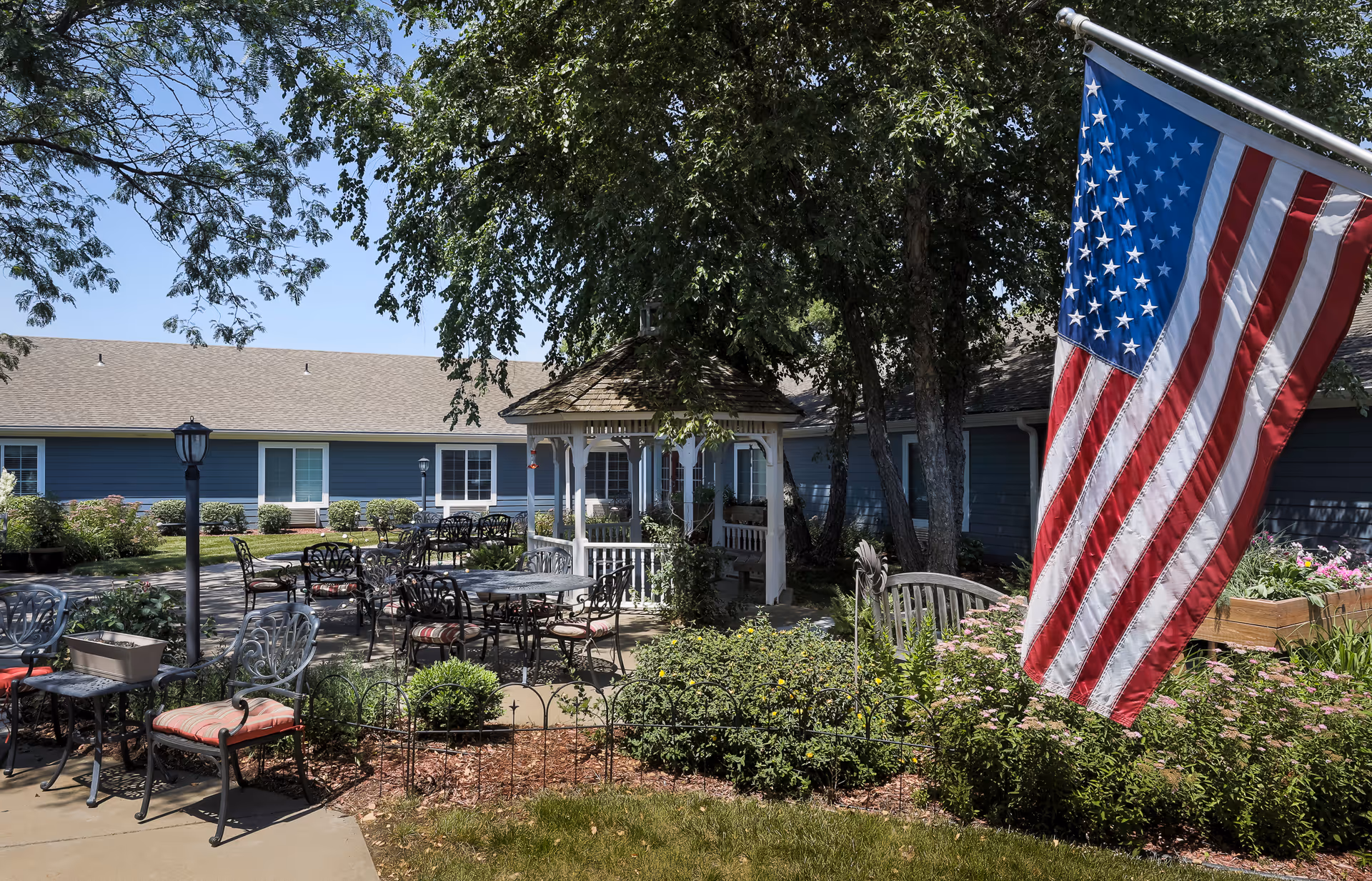 Outdoor courtyard area at Homestead Assisted Living & Memory Care of Topeka featuring metal tables and chairs with cushions, a white gazebo surrounded by trees and shrubs, and an American flag prominently displayed on the right side. The building with blue siding and white-trimmed windows is visible in the background under a clear blue sky.